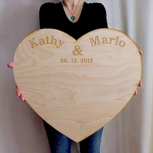 K&ouml;nnte beinhalten: Ein gro&szlig;es, herzf&ouml;rmiges Holzschild mit den Namen "Kathy & Mario" und dem Datum "06. 12. 2015" in goldenen Lettern graviert.