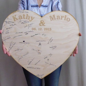 K&ouml;nnte beinhalten: Ein herzf&ouml;rmiges Holzschild mit den Namen "Kathy & Mario" und dem Datum "06.12.2015" eingraviert. Das Schild ist mit Unterschriften und Zeichnungen bedeckt.