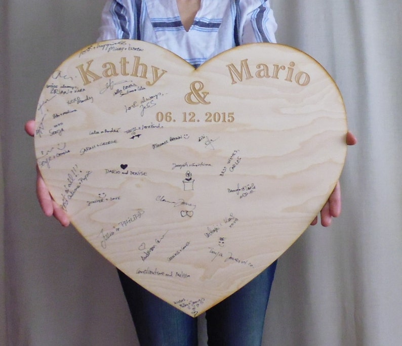 K&ouml;nnte beinhalten: Ein herzf&ouml;rmiges Holzschild mit den Namen "Kathy & Mario" und dem Datum "06.12.2015" eingraviert. Das Schild ist mit Unterschriften und Zeichnungen bedeckt.