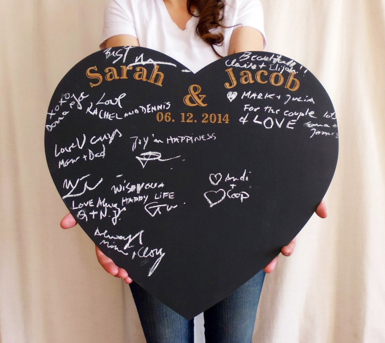 K&ouml;nnte beinhalten: Eine schwarze herzf&ouml;rmige Tafel mit den Namen Sarah und Jacob in goldenen Lettern. Das Datum 06.12.2014 ist unter den Namen geschrieben. Die Tafel ist mit wei&szlig;er Kreide beschriftet, darunter Gl&uuml;ckw&uuml;nsche f&uuml;r das Paar.