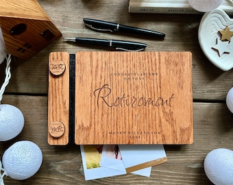 May include: A wooden retirement guest book with the words "Congratulations on Your Retirement" engraved on the front. The book includes the name "Mandy Richardson" and the year "2024". Two black pens and a small wooden house are also visible.