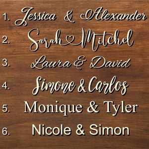 K&ouml;nnte beinhalten: Ein Holzschild mit einer Liste von Namen und Nummern. Die Namen sind in einer kursiven Schrift geschrieben und beinhalten Jessica & Alexander, Sarah Mitchel, Laura & David, Simone & Carlos, Monique & Tyler und Nicole & Simon.