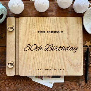 Könnte beinhalten: Ein Fotoalbum aus Holz mit dem eingravierten Text "PETER ROBERTSON'S 80th Birthday" und "EST. JULY 1st, 1945" auf dem Einband. Das Album ist geöffnet und zeigt Fotos im Inneren. Eine hellblaue Sofortbildkamera und dekorative Lichter im Hintergrund.