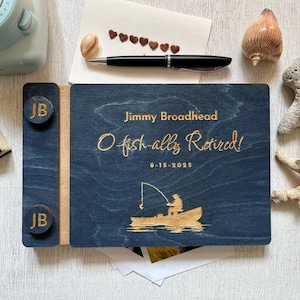 Könnte beinhalten: Ein marineblaues Gästebuch zur Rente aus Holz mit dem Text "Jimmy Broadhead, O fish-ally Retired!" und einer Illustration eines Fischerbootes. Enthält einen Stift, Muscheln und ein kleines Buch. Das Buch misst ca. 20 x 25 cm.