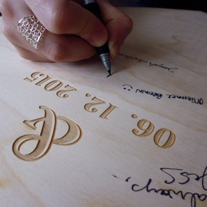 K&ouml;nnte beinhalten: Nahaufnahme eines Holzschildes mit dem Datum "06.12.2012" und dem gravierten Buchstaben "P". Das Schild wird von einer Person mit einem schwarzen Stift signiert.