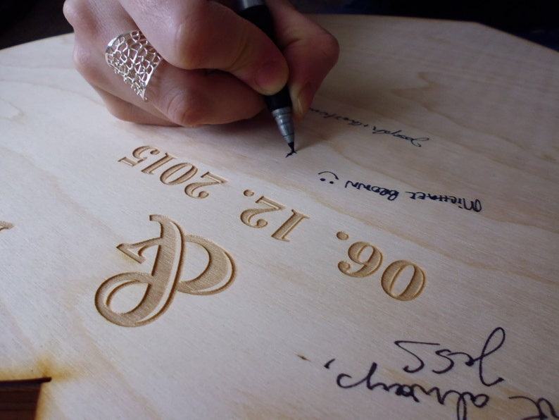 K&ouml;nnte beinhalten: Nahaufnahme eines Holzschildes mit dem Datum "06.12.2012" und dem gravierten Buchstaben "P". Das Schild wird von einer Person mit einem schwarzen Stift signiert.