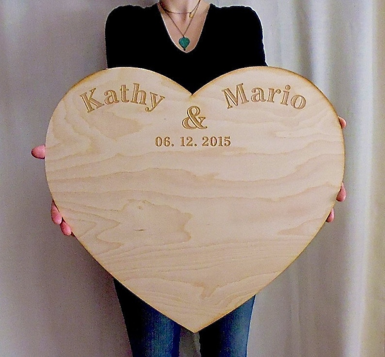 K&ouml;nnte beinhalten: Ein gro&szlig;es, herzf&ouml;rmiges Holzschild mit den Namen "Kathy & Mario" und dem Datum "06. 12. 2015" in goldenen Lettern graviert.
