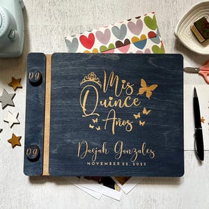 Könnte beinhalten: Ein marineblaues Fotoalbum aus Holz mit goldfarbenem Schriftzug „Mis Quince Años“ und dem Namen „Daejah Gonzales“ mit dem Datum 22. November 2023. Das Album ist von dekorativen Gegenständen umgeben.