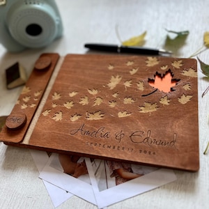 Könnte beinhalten: Ein Fotoalbum aus Holz mit Herbstthema. Der Einband ist mit den Namen "Amelia & Edward" und dem Datum "SEPTEMBER 17, 2024" graviert. Das Album hat einen Ahornblatt-Ausschnitt und Blattdesigns. Ein Stift und eine Sofortbildkamera sind sichtbar.