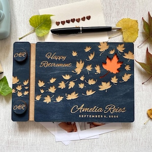 Könnte beinhalten: Eine marineblaue Holz-Geschenkbox zur Rente mit goldenen Blattausschnitten und Text. Auf der Box steht "Happy Retirement" und "Amelia Reis, September 8, 2026". Zwei runde Holzknöpfe mit den Initialen "AR" befinden sich an der Seite.