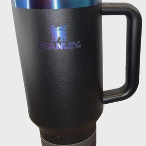 May include: Black Stanley Quencher H Tumbler with a handle and a straw. The tumbler has a black lid with a blue iridescent band. The Stanley logo is in blue. The tumbler holds 40 oz (1.18 L).