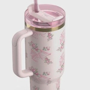 May include: A pink insulated travel mug with a handle, lid, and straw. The mug has a repeating pattern of pink bows and roses on a white background. The lid and handle are also pink, with a gold band near the top.