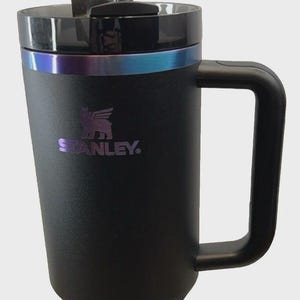 May include: Black Stanley Quencher tumbler with a handle and a straw. The tumbler has a black lid with a metallic iridescent band. The Stanley logo is printed in purple. The tumbler is labeled "Quencher Tumbler" and holds 40 oz.