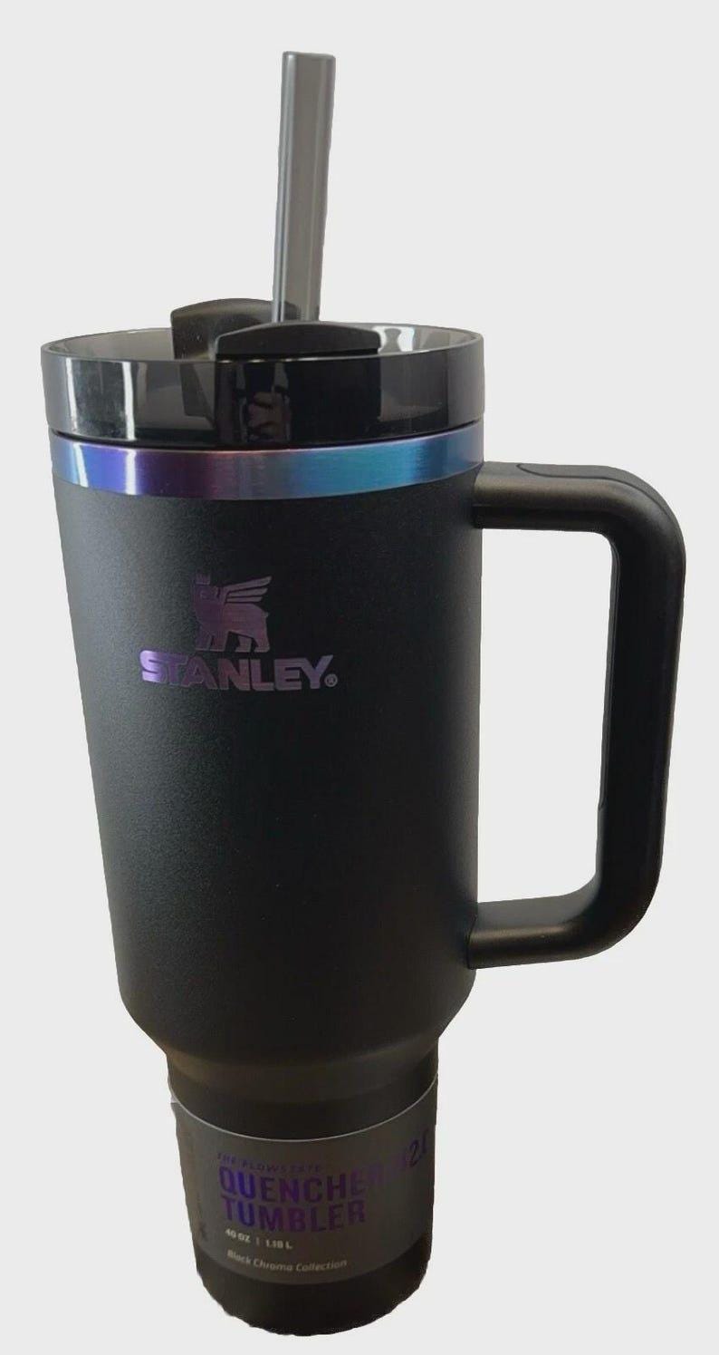 May include: Black Stanley Quencher tumbler with a handle and a straw. The tumbler has a black lid with a metallic iridescent band. The Stanley logo is printed in purple. The tumbler is labeled "Quencher Tumbler" and holds 40 oz.