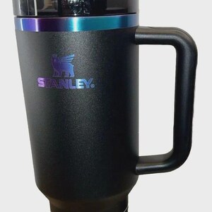 May include: Black Stanley Quencher H2.0 Tumbler with a handle and straw. The tumbler has a black lid with a teal and purple iridescent band. The Stanley logo is visible on the front. The bottom of the tumbler has a label that reads "Quencher H2.0 Tumbler".