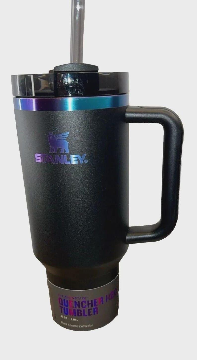 May include: Black Stanley Quencher H2.0 Tumbler with a handle and straw. The tumbler has a black lid with a teal and purple iridescent band. The Stanley logo is visible on the front. The bottom of the tumbler has a label that reads "Quencher H2.0 Tumbler".