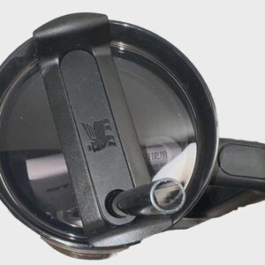 May include: Close-up of a black Stanley tumbler lid with a flip-top closure and a handle. The lid features a black sliding mechanism and a silver logo. The tumbler is designed for hot and cold beverages.
