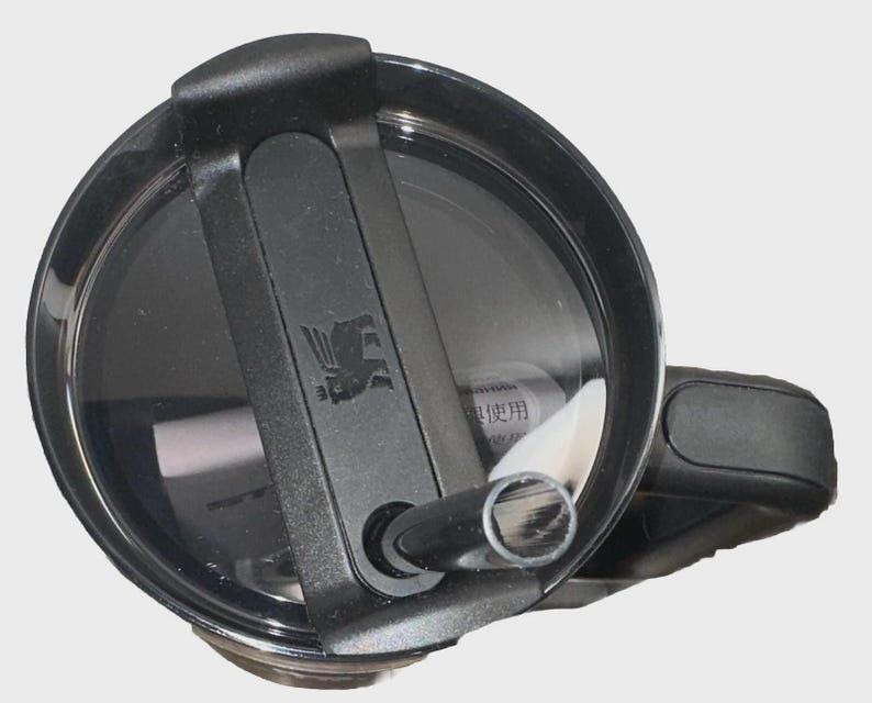 May include: Close-up of a black Stanley tumbler lid with a flip-top closure and a handle. The lid features a black sliding mechanism and a silver logo. The tumbler is designed for hot and cold beverages.