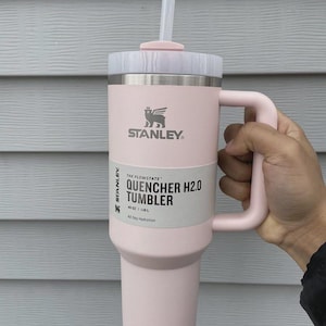 May include: A light pink Stanley Quencher H2.0 Tumbler with a handle and straw. The tumbler has a white label with the text "QUENCHER H2.0 TUMBLER" and "40 OZ | 1.18 L". The Stanley logo is visible.
