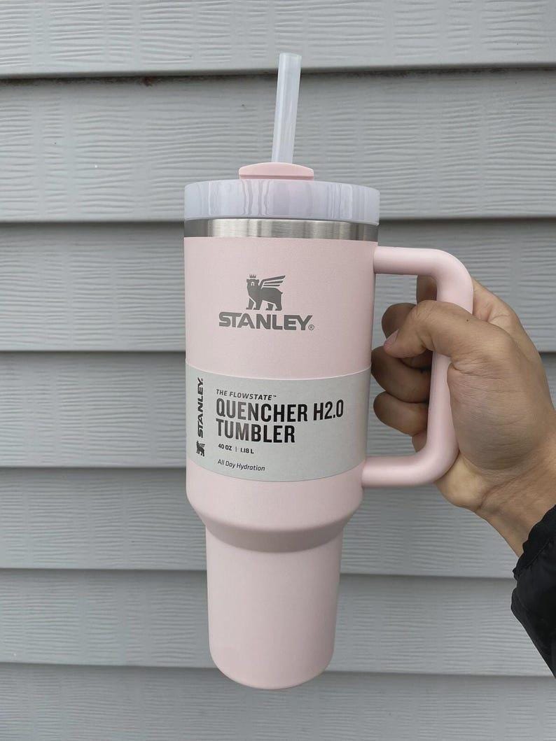 May include: A light pink Stanley Quencher H2.0 Tumbler with a handle and straw. The tumbler has a white label with the text "QUENCHER H2.0 TUMBLER" and "40 OZ | 1.18 L". The Stanley logo is visible.