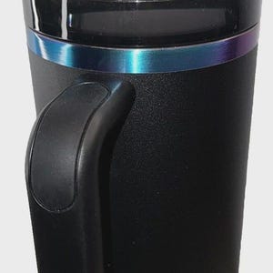 May include: Black insulated tumbler with a handle and a clear lid. The tumbler has a metallic iridescent band around the middle and a clear straw. The bottom of the tumbler has a textured design.