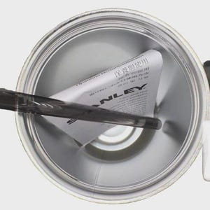 May include: An overhead view of a silver-colored Stanley thermos. A black handle and white plastic component are visible on the right side. A dark gray stirrer is inside the thermos, and a folded paper with text is inside.