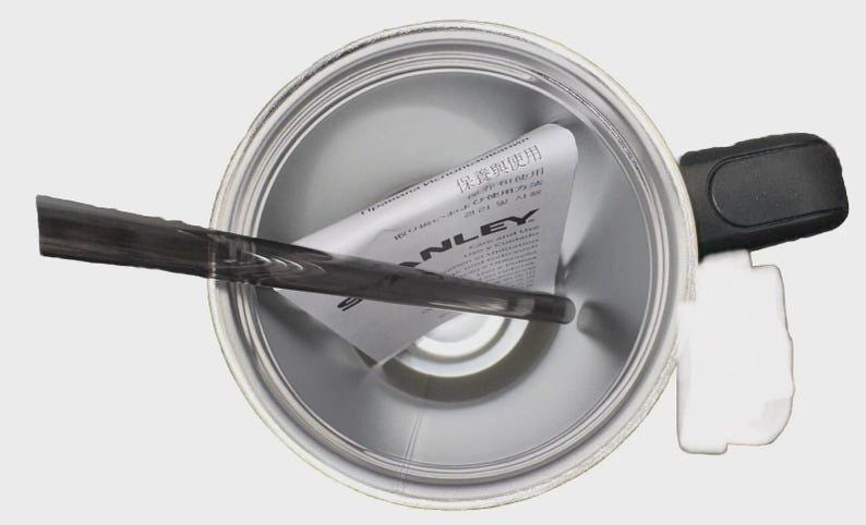 May include: An overhead view of a silver-colored Stanley thermos. A black handle and white plastic component are visible on the right side. A dark gray stirrer is inside the thermos, and a folded paper with text is inside.