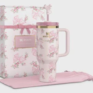 May include: A white Stanley tumbler with a pink floral and bow pattern, a matching handle, and a straw. The tumbler is next to a gift box with the same pattern, a pink ribbon, and a pink drawstring bag.
