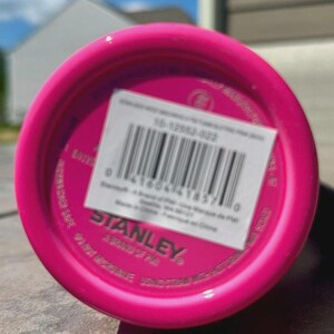 May include: Close-up of the bottom of a bright pink Stanley water bottle. A white sticker with a barcode and product information is affixed to the base. The Stanley logo is visible.