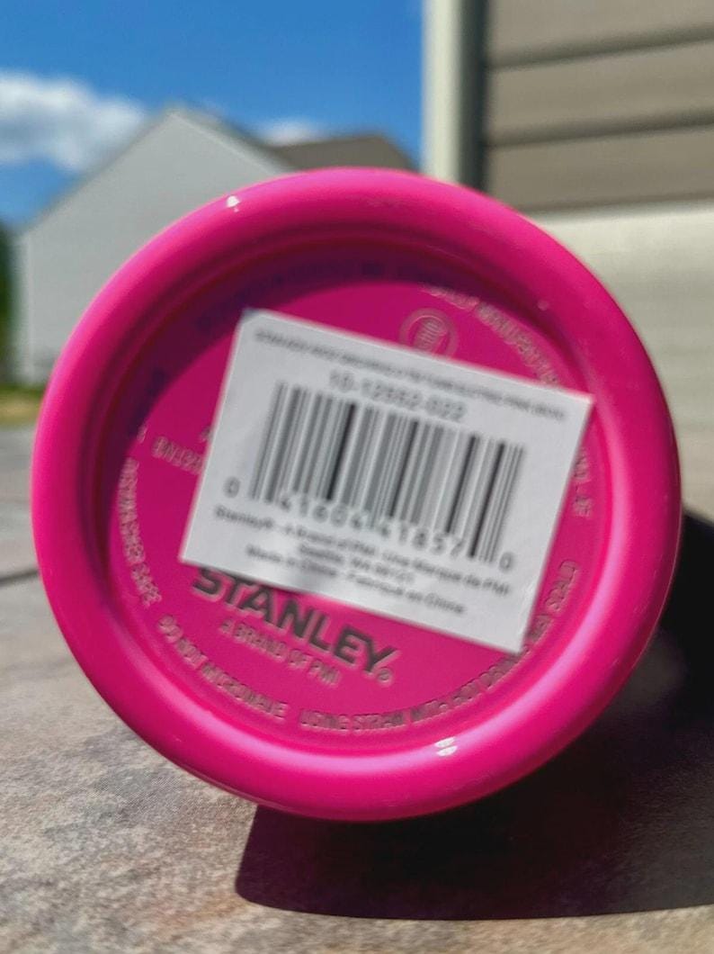 May include: Close-up of the bottom of a bright pink Stanley water bottle. A white sticker with a barcode and product information is affixed to the base. The Stanley logo is visible.