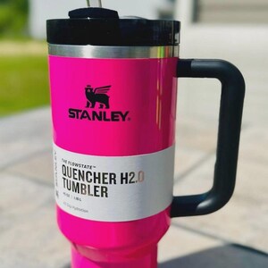 May include: Bright pink Stanley Quencher H2.0 tumbler with a black handle and lid. The tumbler has a clear straw and a white label with the text "QUENCHER H2.0 TUMBLER". The Stanley logo is visible.