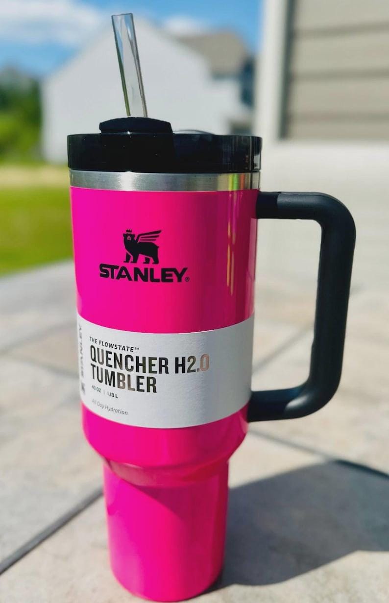May include: Bright pink Stanley Quencher H2.0 tumbler with a black handle and lid. The tumbler has a clear straw and a white label with the text "QUENCHER H2.0 TUMBLER". The Stanley logo is visible.