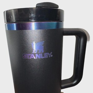 May include: Black Stanley travel mug with a handle and a black lid. The mug has a silver band and a blue and purple iridescent accent. The Stanley logo is printed in blue on the front of the mug.