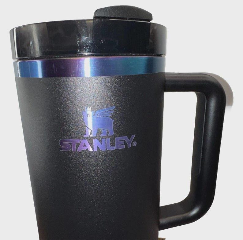 May include: Black Stanley travel mug with a handle and a black lid. The mug has a silver band and a blue and purple iridescent accent. The Stanley logo is printed in blue on the front of the mug.