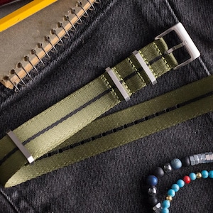 May include: An olive green nylon watch strap featuring a thin black center stripe, a silver-toned metal buckle, and two matching silver-toned metal keepers. The strap has a series of rectangular holes for adjustment and a pointed end.