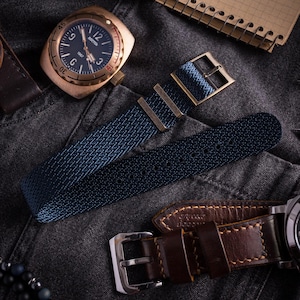 May include: A bronze-toned wristwatch with a black dial featuring the text BOSTOK 1967, paired with a dark blue woven fabric watch strap that has a metal buckle and two metal keepers, alongside a separate dark brown leather watch strap.