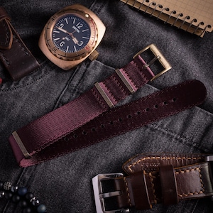 Pode incluir: Um relógio de pulso cor de bronze com mostrador preto exibindo o texto Vostok 1967, acompanhado por uma pulseira de nylon bordô escuro com ferragens de metal bronze e uma pulseira de couro castanho escuro com fivela de metal prateado.