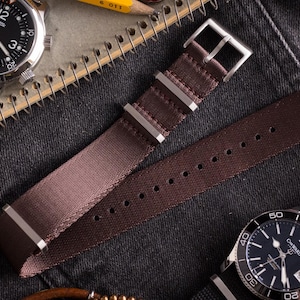 May include: A dark brown nylon watch strap featuring a silver-toned metal buckle and two matching metal keepers. The strap has a woven texture and a series of punched holes for adjustment, presented alongside two analog wristwatches with black dials.