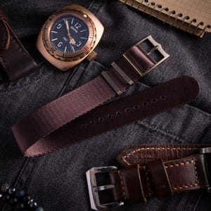 Peut inclure: Une montre-bracelet couleur bronze avec un cadran noir portant l'inscription BOSTOK 1967, accompagnée d'un bracelet en nylon marron foncé avec boucle métallique et d'un bracelet en cuir marron foncé avec coutures et boucle en métal argenté.