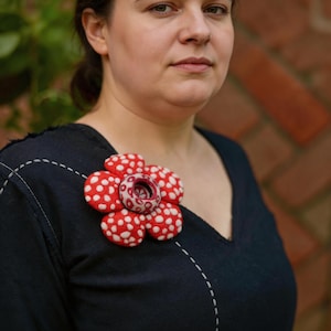 Peut inclure: Une chemise bleu marine à col en V ornée d'une grande broche en forme de fleur à cinq pétales. La broche présente des pétales rouges à pois blancs et un élément central circulaire avec un motif blanc et rouge, épinglée sur la poitrine.