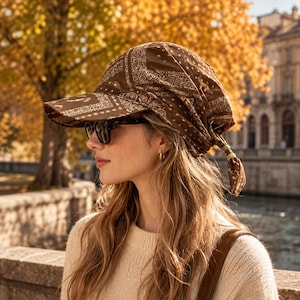 Può includere: Un cappellino da baseball marrone con un motivo a bandana paisley bianco. Il cappellino presenta una visiera strutturata e una corona in tessuto morbido che si raccoglie sul retro, fissata con un piccolo nodo. Il design incorpora motivi a bandana.