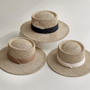 May include: Three straw hats with wide brims and flat crowns. Each hat features a woven texture and a fabric ribbon band around the base of the crown. The ribbons are colored black, tan, and white, respectively, providing a distinct contrast.