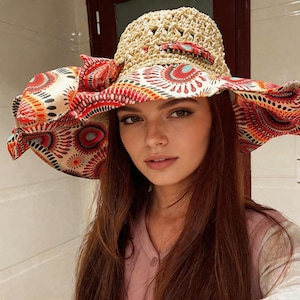 May include: A wide-brimmed sun hat featuring a beige crocheted crown and a large, floppy brim made of fabric with a vibrant, multicolored circular pattern in shades of red, orange, and teal. The hat includes a decorative fabric bow on the side.