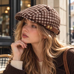May include: A brown and white checkered flat cap featuring a structured brim and a side strap detail secured with two metallic gold-toned buttons. The cap is crafted from a woven fabric with a classic plaid pattern and a rounded, fitted crown shape.