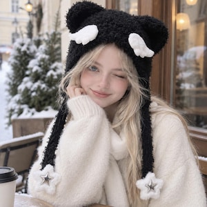 May include: A black fuzzy beanie featuring two rounded ears, two small white wings attached to the sides, and two long dangling earflaps. Each earflap ends in a white plush star with a metallic silver star emblem centered on the front surface.