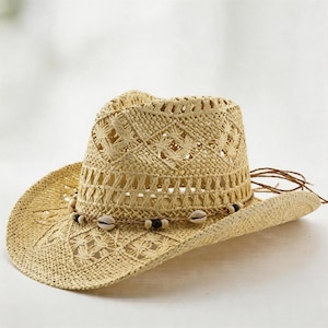 May include: A light beige straw hat featuring an open-weave diamond pattern throughout the crown and brim. The hat is accented with a thin cord band decorated with small white seashells and dark brown beads, with loose cord ends at the back.