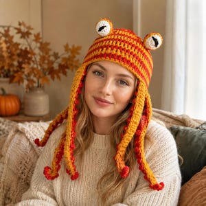 May include: A crochet hat featuring a striped orange and yellow pattern with two protruding eyes on top and long, dangling side panels. The hat is constructed with a textured yarn finish, incorporating small red accents along the edges of the side panels.