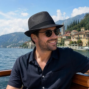 May include: A black straw fedora hat with a solid black ribbon band, paired with dark-tinted sunglasses and a navy blue short-sleeved button-down shirt. The hat features a woven texture and a structured crown with a slightly upturned brim.