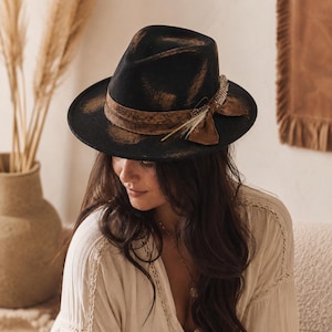 Könnte beinhalten: Ein schwarzer Fedora-Hut mit einem abgenutzten braunen Finish auf der Krone und der Krempe. Der Hut ist mit einem breiten braunen Band akzentuiert, das an der Seite zu einer Schleife gebunden und mit einer kleinen hellen Feder verziert ist.