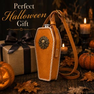 May include: A tan leather coffin-shaped flask holder with white cross-stitching, featuring a metallic skull and clock face emblem on the front. It includes an adjustable leather shoulder strap and a silver-toned metal cap at the top.